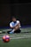 A man sits on a sports field, wearing a blue sports vest over a white shirt and black shorts. His legs are crossed, and he appears to be resting or taking a break. The background is dimly lit, with a blurred out soccer ball in the foreground and netting visible behind him.
