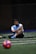 A man sits on a sports field, wearing a blue sports vest over a white shirt and black shorts. His legs are crossed, and he appears to be resting or taking a break. The background is dimly lit, with a blurred out soccer ball in the foreground and netting visible behind him.