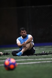 A man sits on a sports field, wearing a blue sports vest over a white shirt and black shorts. His legs are crossed, and he appears to be resting or taking a break. The background is dimly lit, with a blurred out soccer ball in the foreground and netting visible behind him.