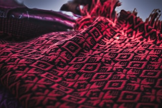 A close-up of a red and black textile with intricate geometric patterns. The fabric features diamond shapes and line motifs, providing a textured and woven appearance. The edges are adorned with fringes, adding a decorative element.