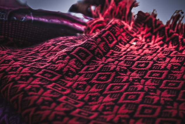 A close-up of a red and black textile with intricate geometric patterns. The fabric features diamond shapes and line motifs, providing a textured and woven appearance. The edges are adorned with fringes, adding a decorative element.