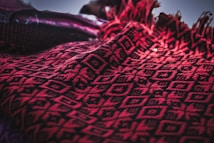 A close-up of a red and black textile with intricate geometric patterns. The fabric features diamond shapes and line motifs, providing a textured and woven appearance. The edges are adorned with fringes, adding a decorative element.