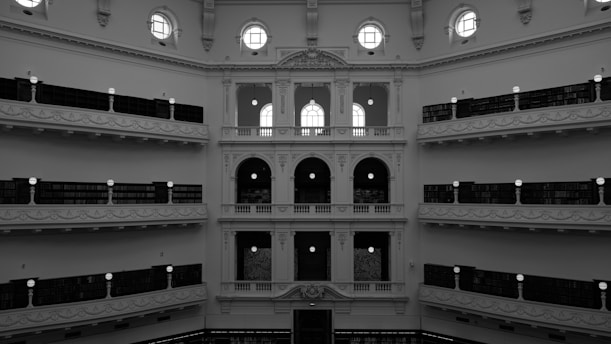 A grand interior of a multi-level library with ornate balconies and archways. Tall shelves filled with books line the walls, and circular windows allow natural light to illuminate the space. The architecture features intricate details and a symmetrical design.