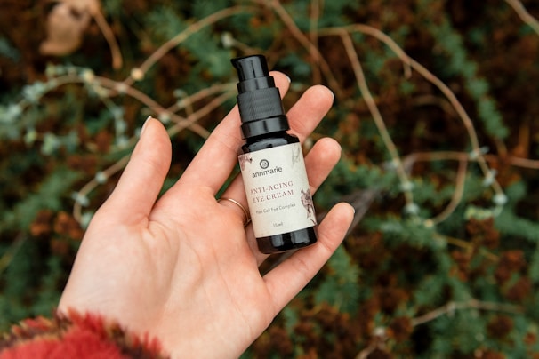A bottle of anti-aging eye cream is held in a person's hand. The background consists of green foliage with dark brownish areas, giving a natural setting.