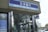 The image depicts the front entrance of a bank with a sign labeled 'North Pacific Bank' in both English and Japanese. The building features mirrored pillars and glass doors. Cars are parked outside, and the background includes other buildings visible in daylight.