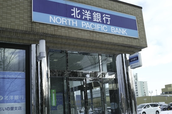The image depicts the front entrance of a bank with a sign labeled 'North Pacific Bank' in both English and Japanese. The building features mirrored pillars and glass doors. Cars are parked outside, and the background includes other buildings visible in daylight.