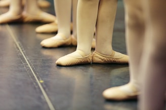 Several ballet dancers stand in a line, focusing on their feet clad in soft ballet slippers. The dancers are aligned on a smooth studio floor, possibly in preparation for a class or rehearsal.