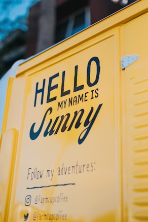 A yellow van with text written on it, reading 'HELLO MY NAME IS Sunny' followed by 'Follow my adventures:' and social media handles. The image is taken at an angle showing part of the side of the van.