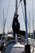 A sailboat is docked in a harbor with its mast and rigging visible. Several other boats are moored nearby, and the water reflects the sunlight.