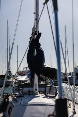 A sailboat is docked in a harbor with its mast and rigging visible. Several other boats are moored nearby, and the water reflects the sunlight.