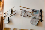 Several newspapers are placed on wooden rods attached to a white wall, creating a simple display. The setup is likely in a waiting area or communal space. Wooden chairs are positioned below the newspapers and part of a wooden shelf with a potted plant is visible on the left.