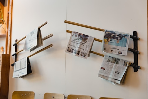 Several newspapers are placed on wooden rods attached to a white wall, creating a simple display. The setup is likely in a waiting area or communal space. Wooden chairs are positioned below the newspapers and part of a wooden shelf with a potted plant is visible on the left.