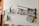 Several newspapers are placed on wooden rods attached to a white wall, creating a simple display. The setup is likely in a waiting area or communal space. Wooden chairs are positioned below the newspapers and part of a wooden shelf with a potted plant is visible on the left.