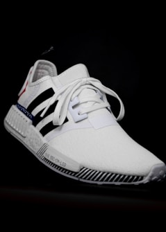 A white athletic shoe with a sleek design, featuring black stripes and a red accent on the heel. The shoe has a textured sole with black and white stripes and white laces tied in the center. The background is black, highlighting the features of the shoe.