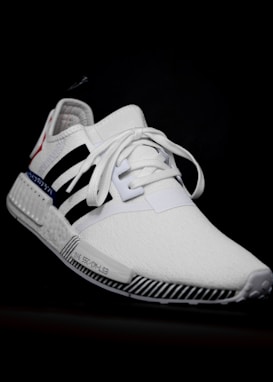 A white athletic shoe with a sleek design, featuring black stripes and a red accent on the heel. The shoe has a textured sole with black and white stripes and white laces tied in the center. The background is black, highlighting the features of the shoe.