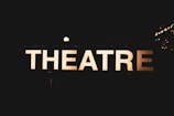 An illuminated sign displaying the word 'THEATRE' against a dark background, creating a contrast that makes the letters stand out sharply. Small bright lights can be seen in the background, suggesting a nighttime or indoor setting.