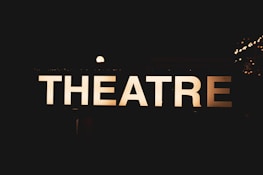 An illuminated sign displaying the word 'THEATRE' against a dark background, creating a contrast that makes the letters stand out sharply. Small bright lights can be seen in the background, suggesting a nighttime or indoor setting.