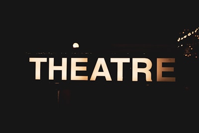 An illuminated sign displaying the word 'THEATRE' against a dark background, creating a contrast that makes the letters stand out sharply. Small bright lights can be seen in the background, suggesting a nighttime or indoor setting.
