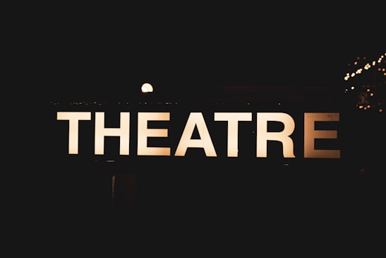 An illuminated sign displaying the word 'THEATRE' against a dark background, creating a contrast that makes the letters stand out sharply. Small bright lights can be seen in the background, suggesting a nighttime or indoor setting.