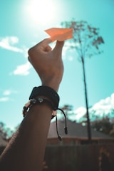 A hand is holding up an orange paper airplane against a bright blue sky with some clouds and a tree in the background. The wrist has a black watch and colorful braided bracelets.