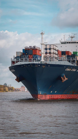 A large container ship moving through a river past a distant city shoreline