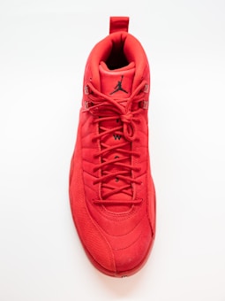 A bright red sneaker viewed from the top. The shoe features a black Jumpman logo on the tongue and detailed stitching throughout. The laces are tightly tied, and the texture includes smooth and textured sections.