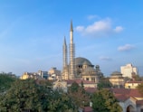 A large mosque with multiple domes and two minarets surrounded by scaffolding. The mosque is set against a clear blue sky with scattered clouds. In the background, various urban buildings can be seen, and in the foreground, there are green trees and red-roofed structures.