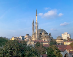 A large mosque with multiple domes and two minarets surrounded by scaffolding. The mosque is set against a clear blue sky with scattered clouds. In the background, various urban buildings can be seen, and in the foreground, there are green trees and red-roofed structures.