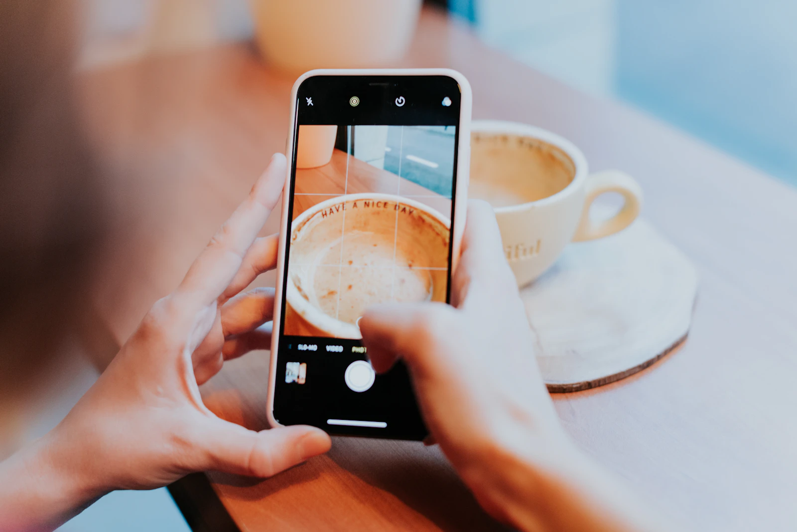 Two hands photographing a cup of coffee with a phone on a bright table