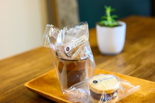 A presentation of packaged snacks on a wooden tray. The packages contain a mini cup cake labeled 'an kurumi' and a cookie labeled 'an butter.' In the background, a small potted plant in a white pot adds a touch of greenery.