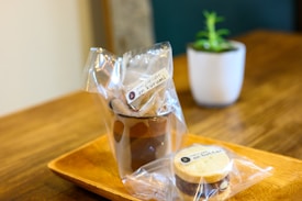 A presentation of packaged snacks on a wooden tray. The packages contain a mini cup cake labeled 'an kurumi' and a cookie labeled 'an butter.' In the background, a small potted plant in a white pot adds a touch of greenery.