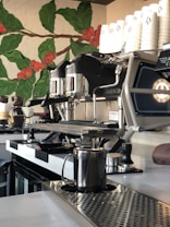 A modern coffee shop counter featuring a sleek espresso machine, a stack of white cups, and various coffee-making tools. Behind the counter, there's a decorative mural of coffee plants with green leaves and ripe red coffee cherries.