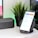 A modern workspace setup featuring a smartphone on a wireless charging stand, displaying a home automation app interface. Next to it is a black portable speaker, and a potted plant adds a touch of greenery. A computer monitor is partially visible in the background.