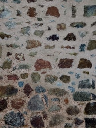 A textured stone wall composed of variously sized and colored stones set in grayish mortar. The stones are irregular in shape, featuring shades of brown, black, green, and gray, creating a rough and rustic appearance.