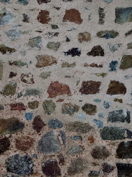A textured stone wall composed of variously sized and colored stones set in grayish mortar. The stones are irregular in shape, featuring shades of brown, black, green, and gray, creating a rough and rustic appearance.