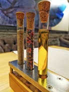 Three glass test tubes are displayed upright in a wooden and metal holder. Each tube has a cork stopper. The left tube contains a coarse sand-like substance, the middle tube is filled with mixed peppercorns, and the right tube contains a yellow liquid.