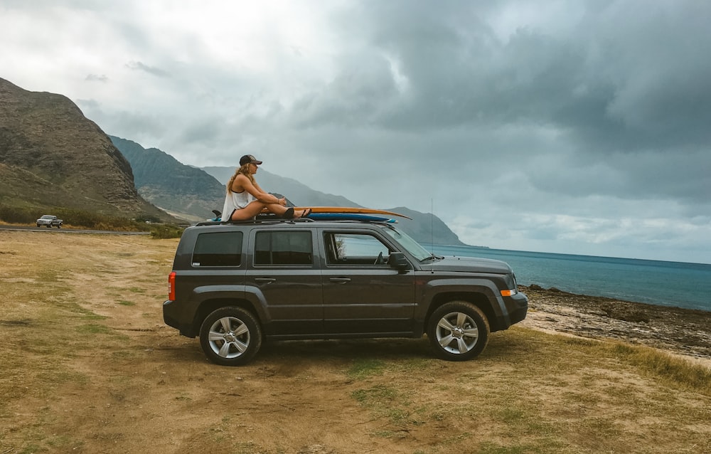 woman wearing white tank top sitting on top of gray SUV viewing body of water and mountain under white and gray sky