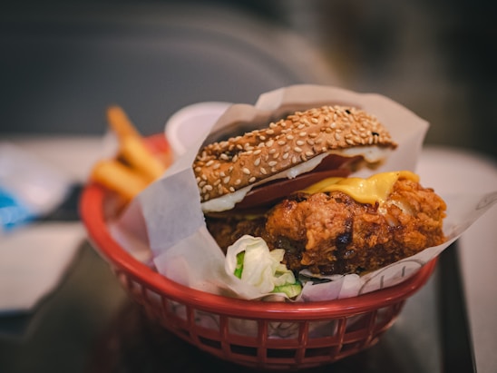 A close-up view of a sesame seed hamburger bun filled with crispy fried chicken, melted cheese, lettuce, and possibly a slice of tomato. The burger is presented in a red basket lined with parchment paper. In the background, blurred french fries and a small container of dipping sauce are visible.