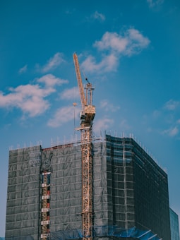 A construction site featuring a tall tower crane is positioned beside a building covered with protective netting. The crane's arm extends upwards against a bright blue sky dotted with clouds.