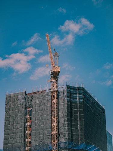 A construction site featuring a tall tower crane is positioned beside a building covered with protective netting. The crane's arm extends upwards against a bright blue sky dotted with clouds.