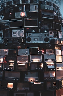 An artistic installation composed of stacked vintage radios of various shapes and sizes. Some radios are illuminated, casting a warm glow, while others remain dark. The collection creates a unique visual texture with metallic and wooden surfaces.