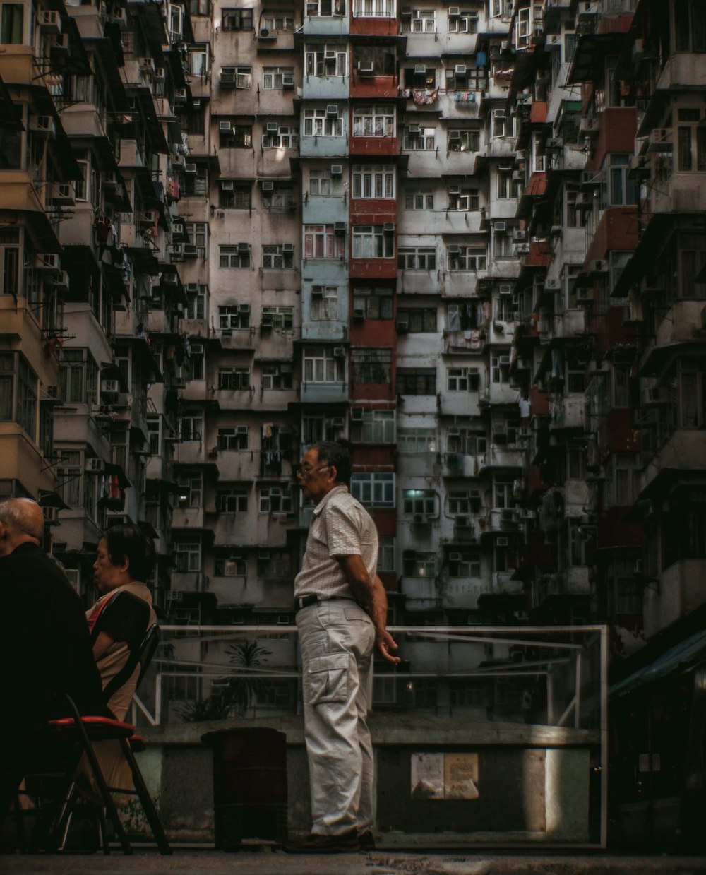 man wearing brown polo shirt standing at the back of people sitting on individual chairs near high-rise buildings