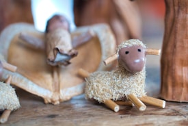 A handcrafted scene depicting a small, rustic model of a lamb with a textured body made of straw-like material and wooden limbs. In the background, there is a figure draped in a fabric, possibly representing an angel or religious figure. The setting appears to be part of a nativity scene, with natural materials used throughout.