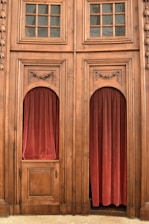 A detailed wooden structure with two large, ornately carved panels featuring glass-paned windows above. Each panel has an alcove filled with red velvet curtains. The wood appears to be aged with intricate designs at the top of the alcoves, giving a classic, vintage look.