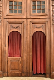 A detailed wooden structure with two large, ornately carved panels featuring glass-paned windows above. Each panel has an alcove filled with red velvet curtains. The wood appears to be aged with intricate designs at the top of the alcoves, giving a classic, vintage look.