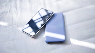 A smartphone with a reflective screen is placed next to a light blue phone case on a white bed sheet. Sunlight is casting soft shadows and highlights across the surface.