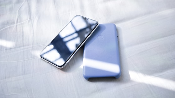 A smartphone with a reflective screen is placed next to a light blue phone case on a white bed sheet. Sunlight is casting soft shadows and highlights across the surface.