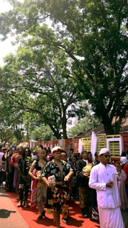 A group of people in traditional attire are participating in a procession under the shade of large trees. The participants appear to be involved in a cultural or ceremonial event, with some holding masks and decorative items.