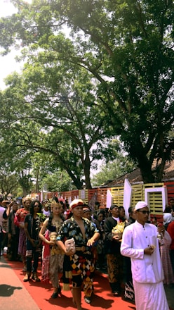 A group of people in traditional attire are participating in a procession under the shade of large trees. The participants appear to be involved in a cultural or ceremonial event, with some holding masks and decorative items.