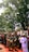 A group of people in traditional attire are participating in a procession under the shade of large trees. The participants appear to be involved in a cultural or ceremonial event, with some holding masks and decorative items.